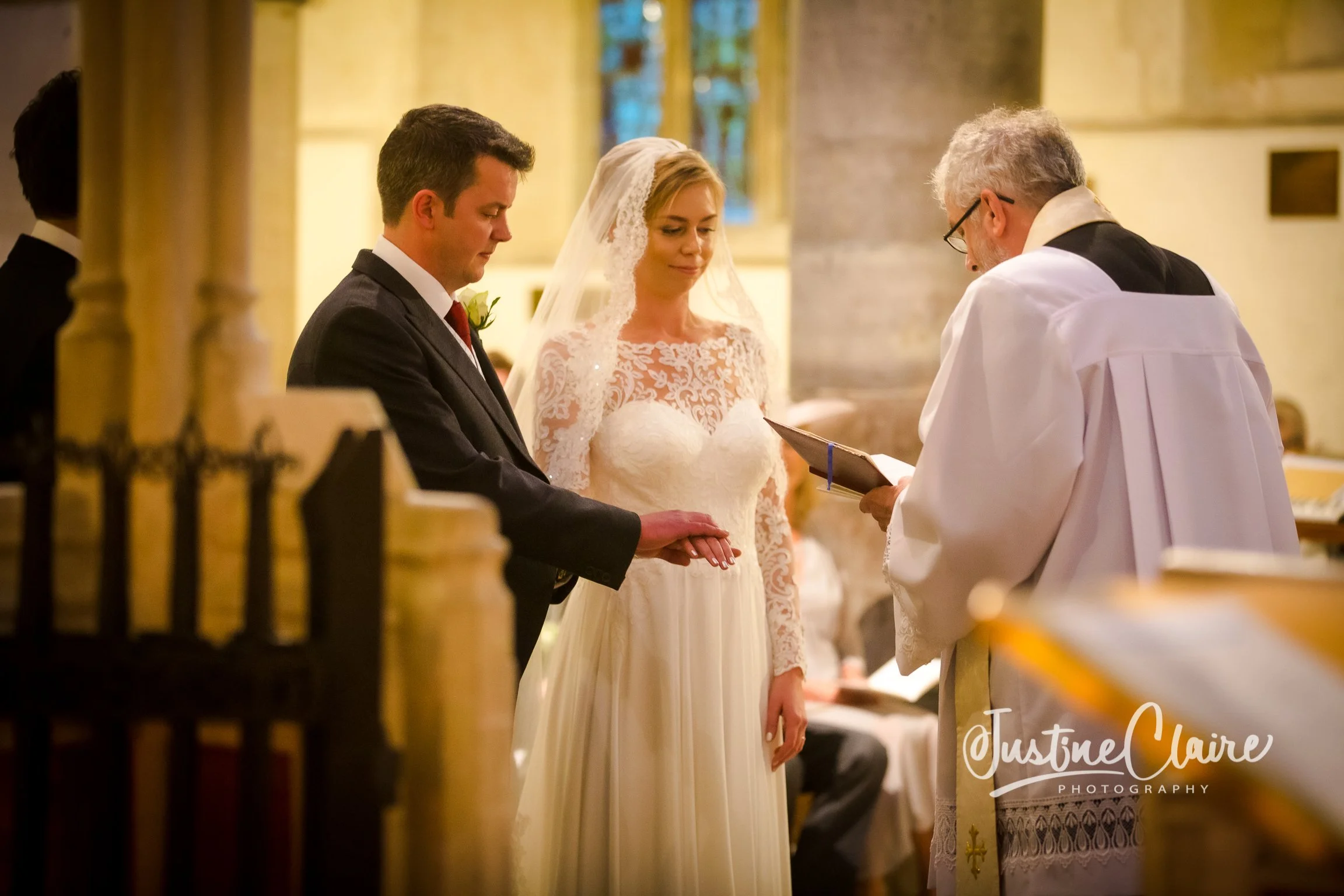 A bride and groom kneeling during a wedding ceremony in a church, with a priest officiating, in front of stained glass windows.