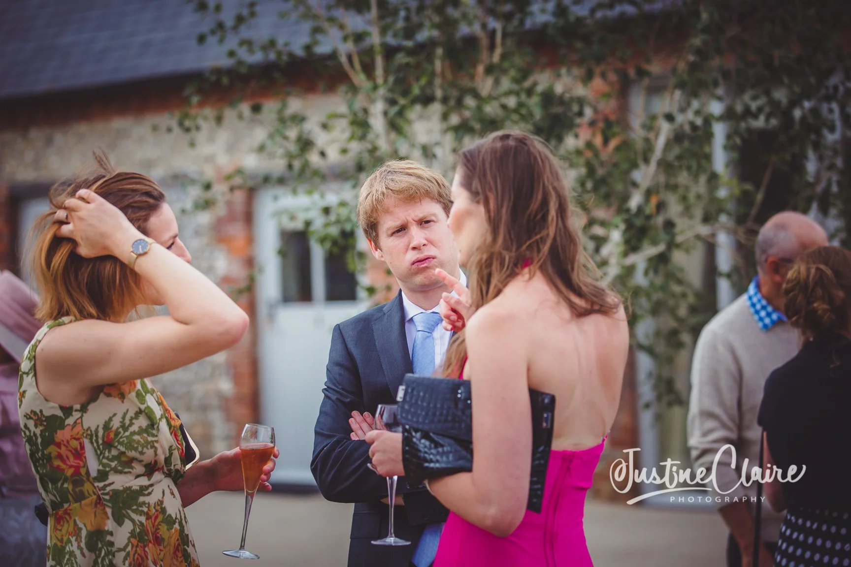 A group of people attending an outdoor social event, including a woman in a floral dress, a man in a suit, and a woman in a pink dress, engaging in conversation with drinks in hand, with a brick house and trees in the background.