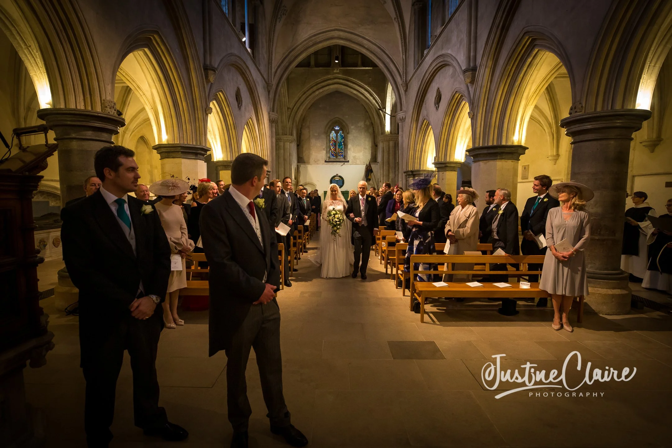 Wedding ceremony inside a historic church with arched ceilings, where the bride is walking down the aisle accompanied by an older man, possibly her father. Guests are standing on both sides of the aisle, dressed in formal attire. The church features