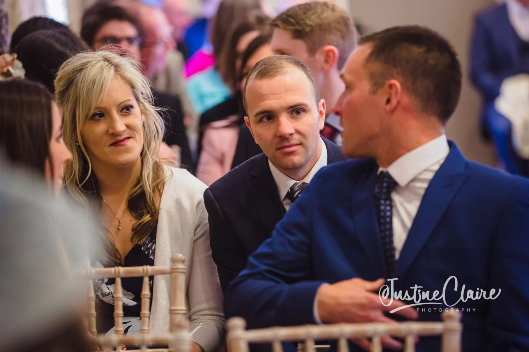 A group of people at a social event, sitting at a table with cream-colored chairs, engaged in conversation. Focus on a woman with long blonde hair and a woman in a blue suit, with other guests in the background.