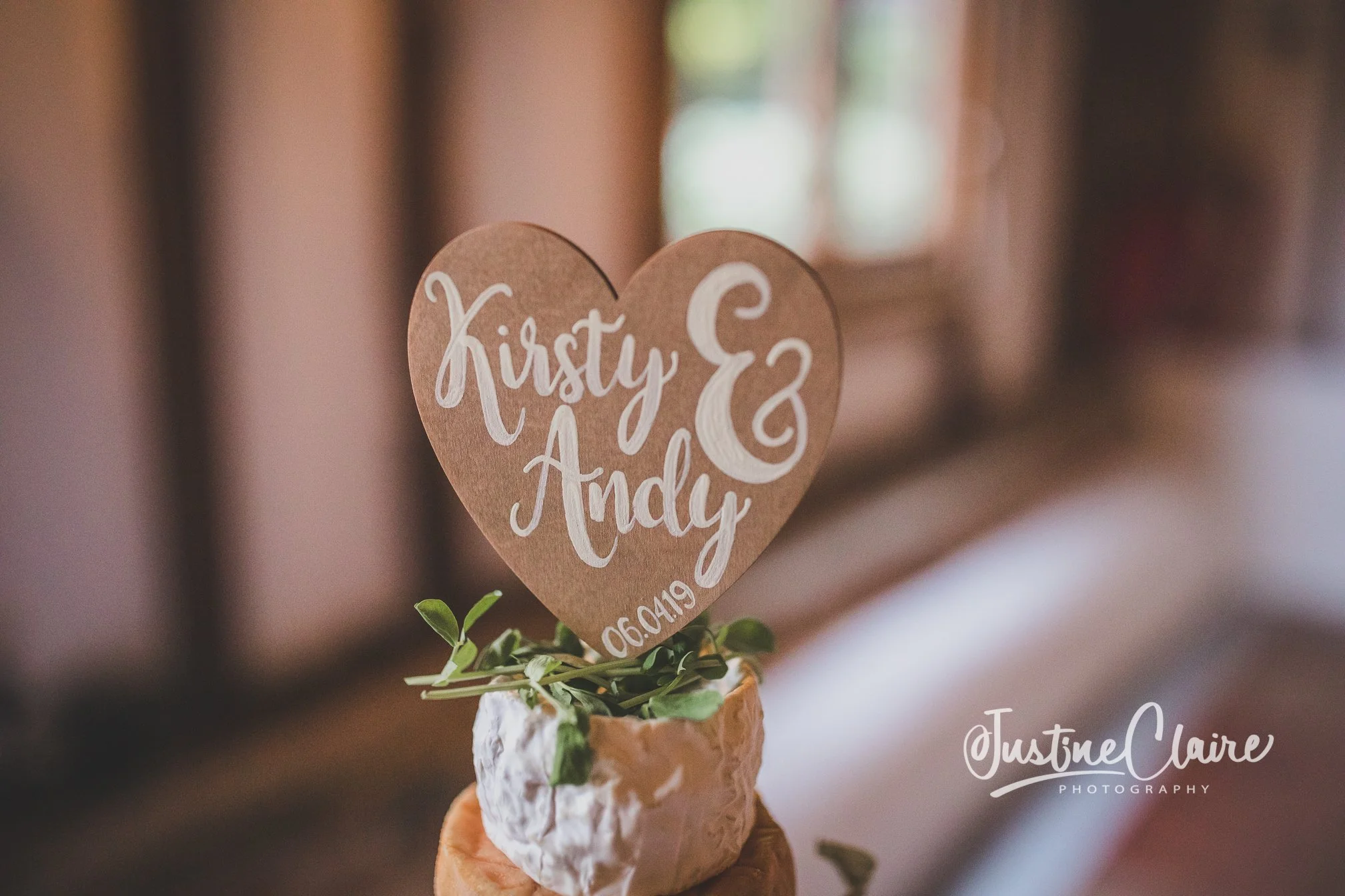 A small plant with green leaves in a white textured pot holds a heart-shaped brown paper sign with white handwritten-style text reading "Kirsty & Andy" and their wedding date "06.04.19". The background is blurred with warm indoor lighting, and a wate