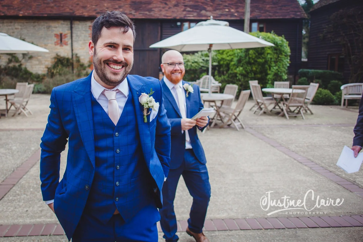 Two men in blue suits, white shirts, and light-colored ties attending an outdoor wedding, with one smiling at the camera in the foreground and the other holding a phone in the background at Fitzleroi barn