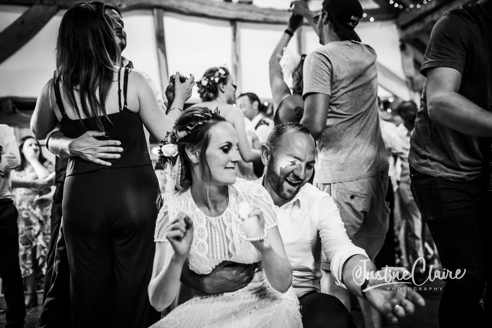 Black and white image of a wedding reception with guests dancing and celebrating, including a woman with a floral crown and lace dress, and a man beside her smiling and celebrating.