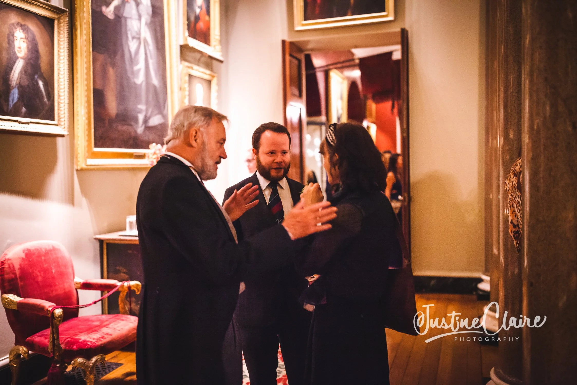 Three people are engaged in a conversation inside an art gallery or museum. The man on the left has white hair and a beard, dressed in a dark suit. The person in the middle, also a man, has dark hair and a beard, wearing a dark suit and striped tie.