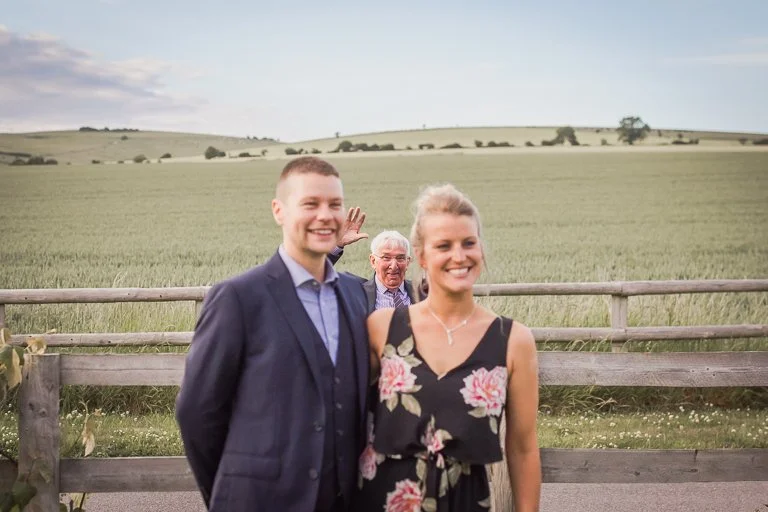 Three people, two women and one man, standing in front of a wooden fence with a rural landscape of rolling green hills and trees in the background, smiling at the camera.