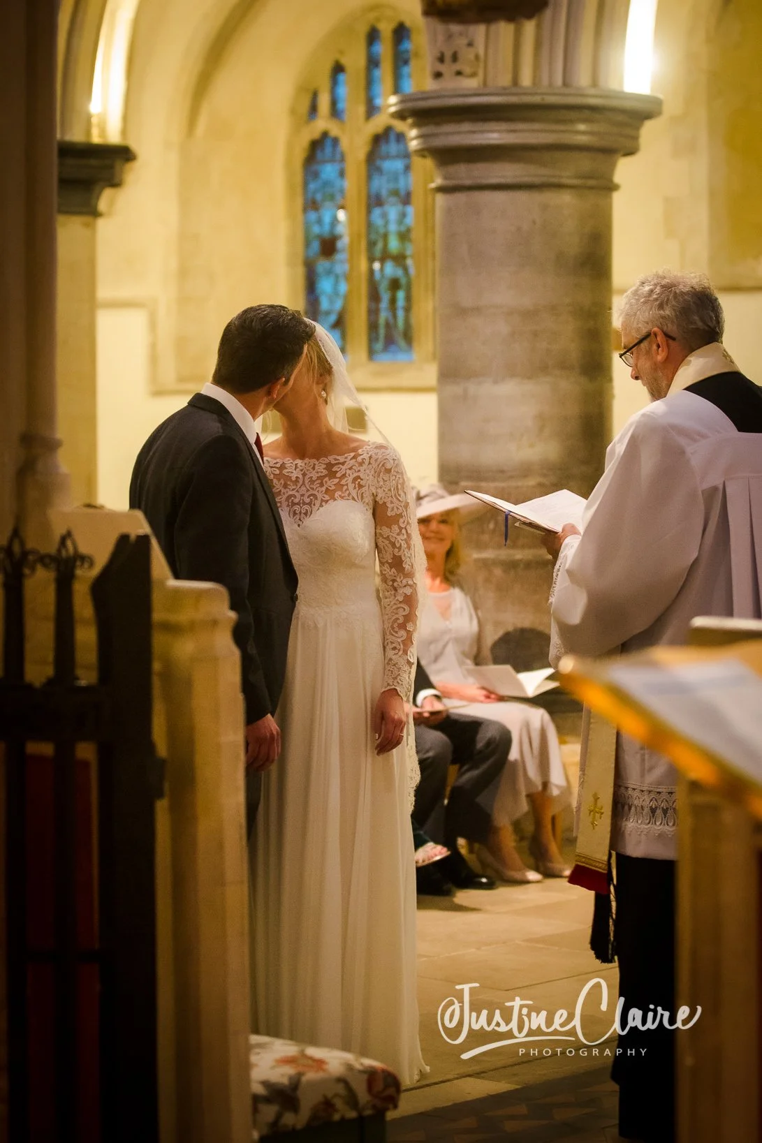 A bride and groom standing before a priest during a wedding ceremony in a church, with seated guests in the background and stained glass windows behind them.