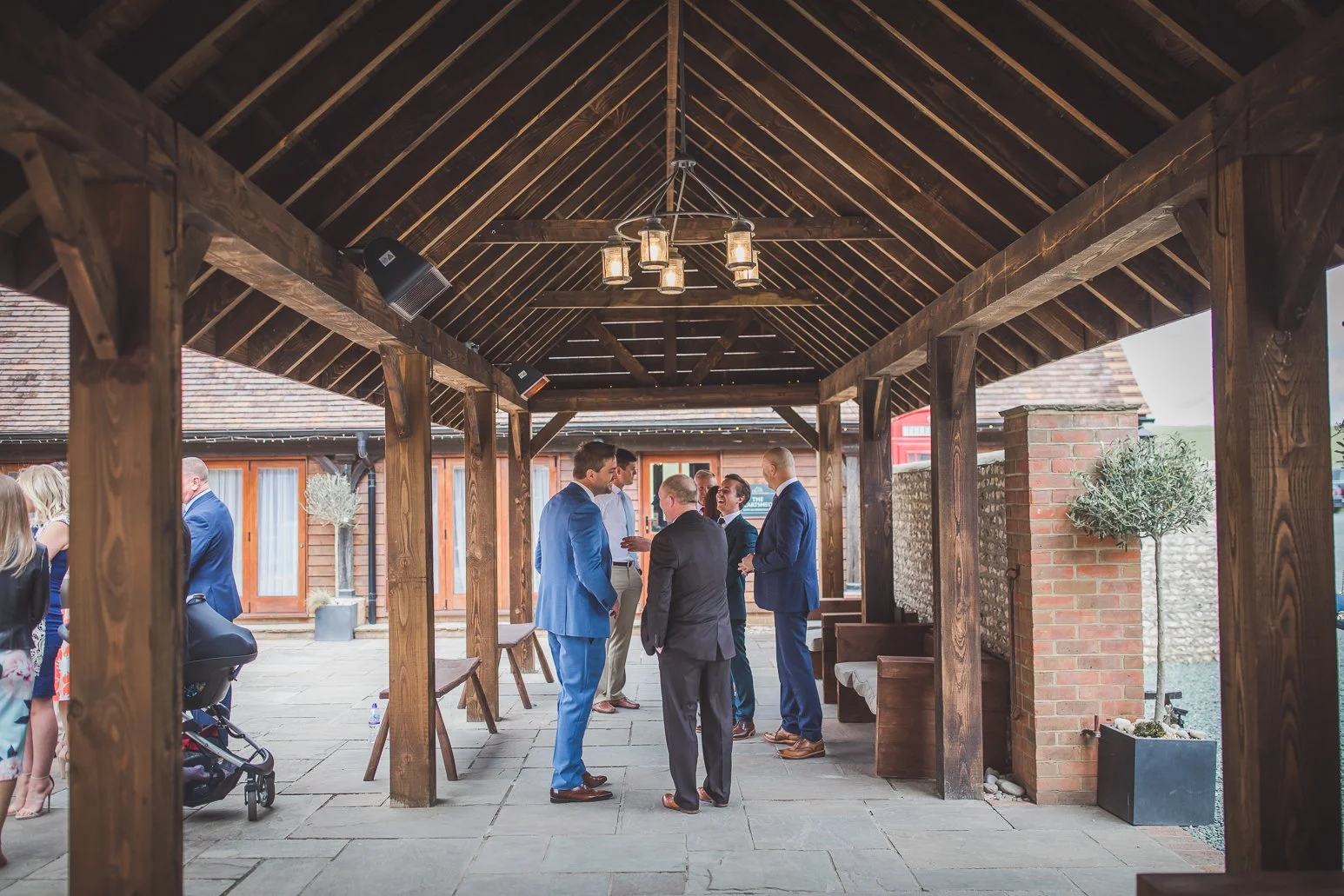 Group of men in suits conversing under a wooden pavilion during a social event, with other guests in the background at an outdoor venue.