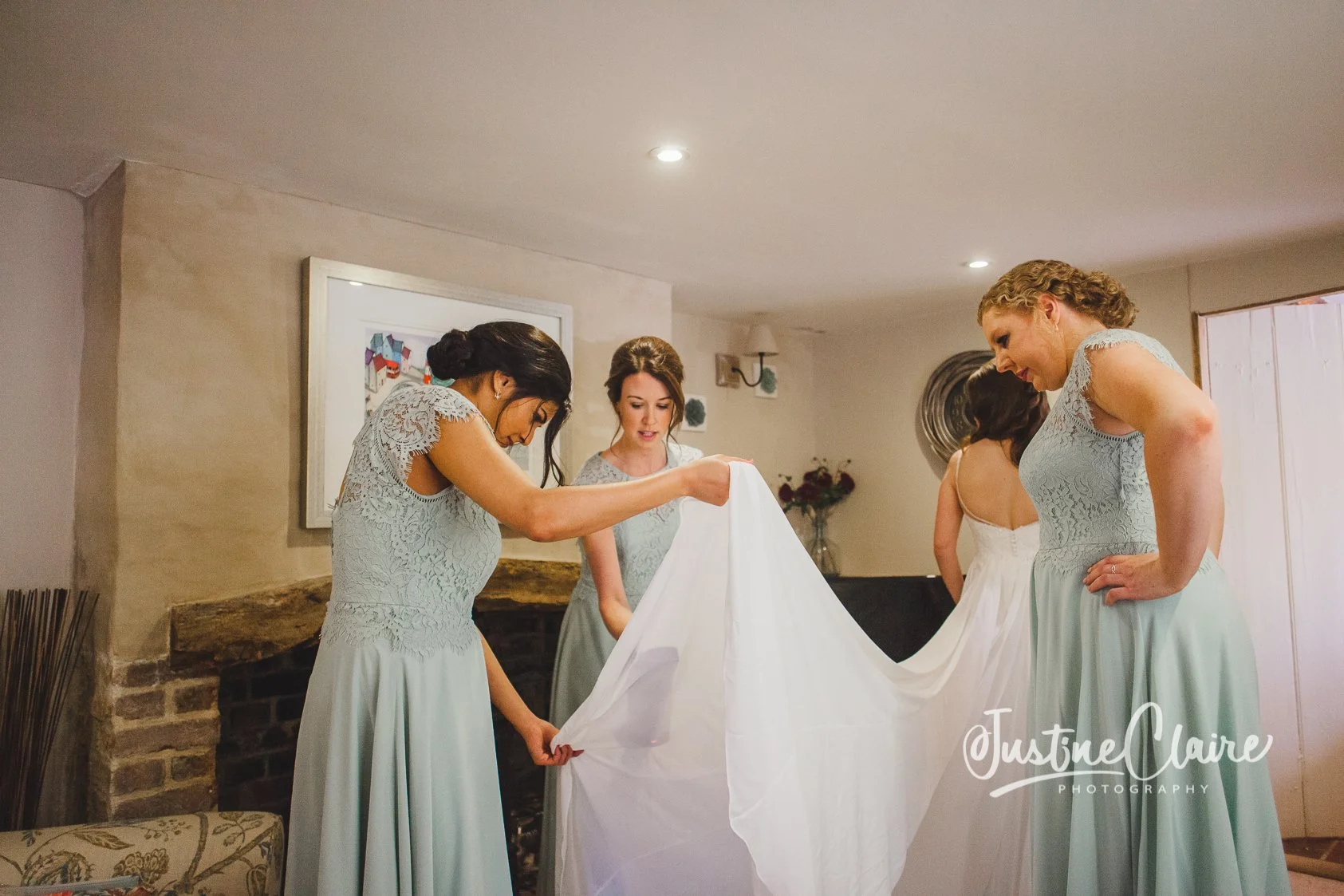 Four women in light blue bridesmaid dresses are gathered around a white wedding dress, preparing it for a wedding in a cozy room in Arundel on Bakers Arms Hill