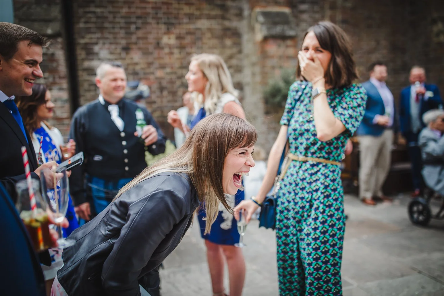 Group of people at a social gathering, with a woman in a leather jacket laughing and a woman in a blue patterned dress covering her mouth, all standing outdoors near a brick wall.