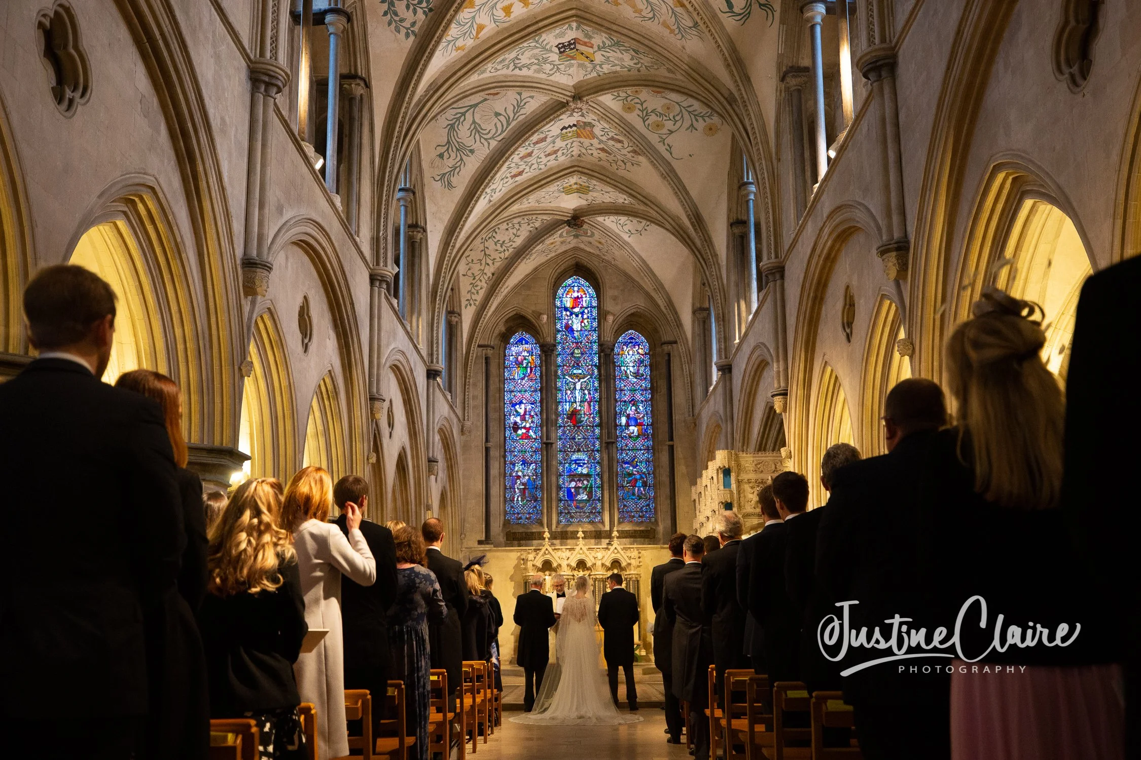 A wedding ceremony inside a grand, historic church with stained glass windows, featuring a bride and groom at the altar with guests seated on either side.