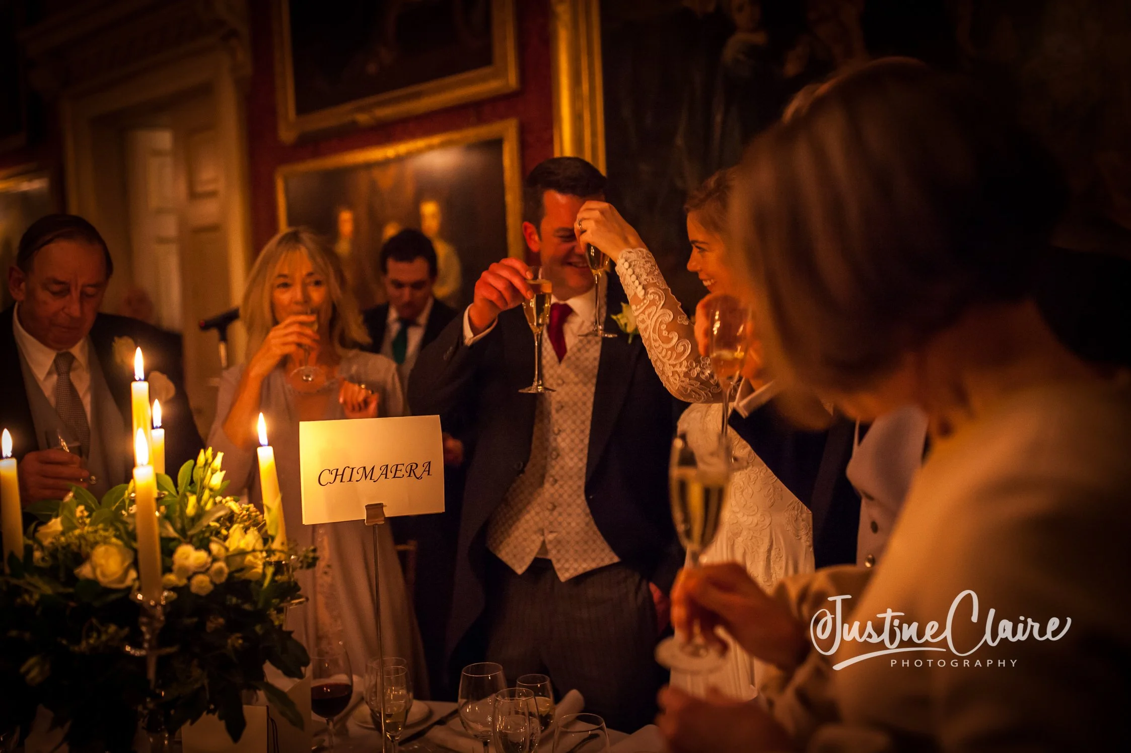 People celebrating at a wedding reception, raising glasses for a toast, in a dimly lit room with candles and floral arrangements.