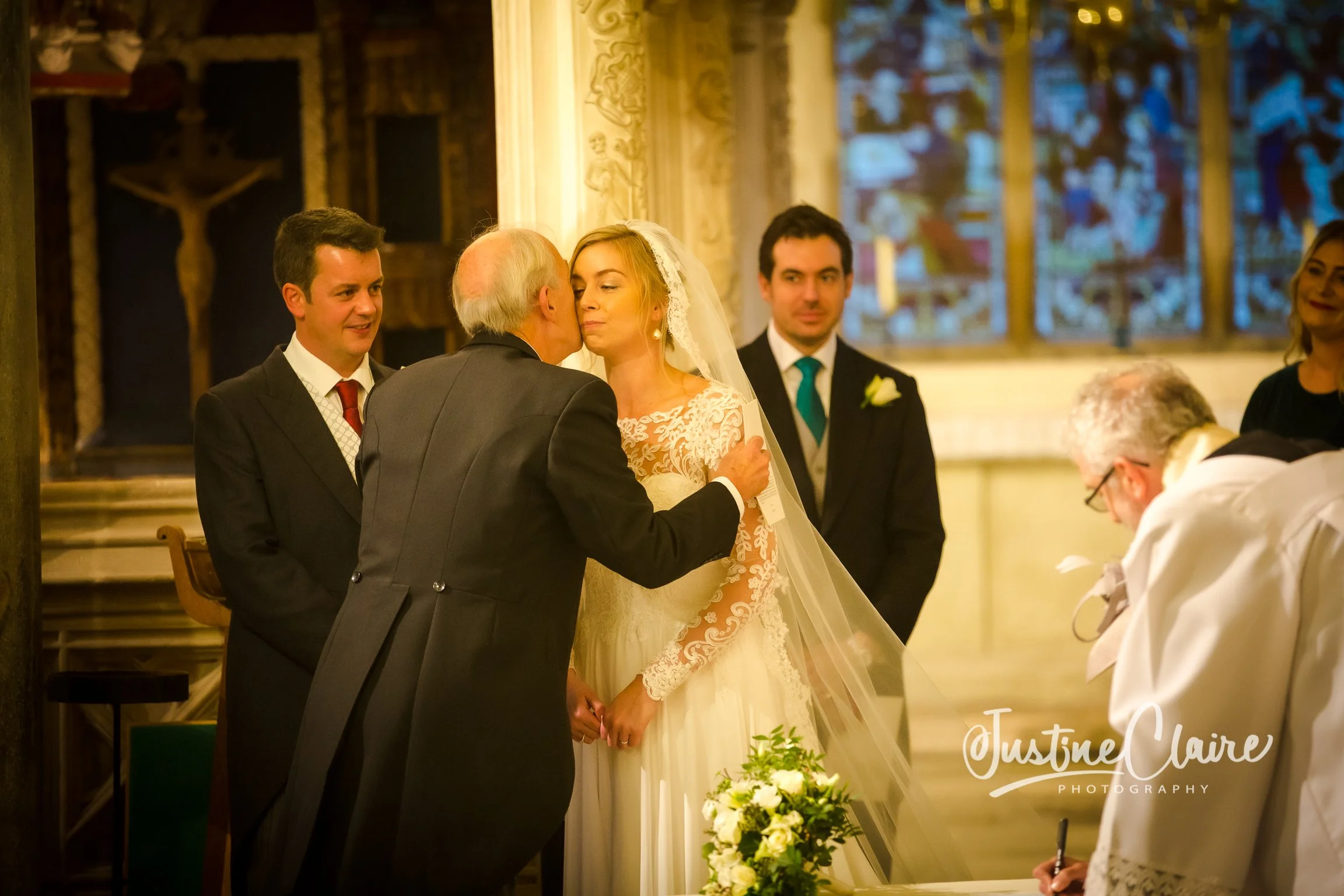 A bride in a lace wedding dress and veil holding hands with an older man, likely her father, in a church. Several men and women in formal attire observe the ceremony, with stained glass windows in the background.