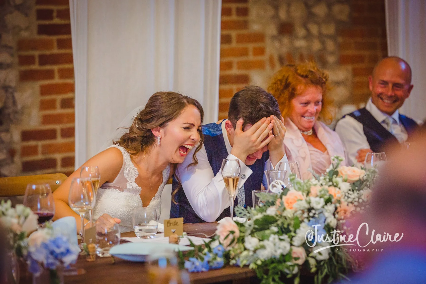 At a wedding reception, a woman in a white dress is laughing with her mouth open, and a man next to her is covering his face with his hands, also laughing. Two other people, a woman with red hair and a man with a bald head, are smiling in the backgro