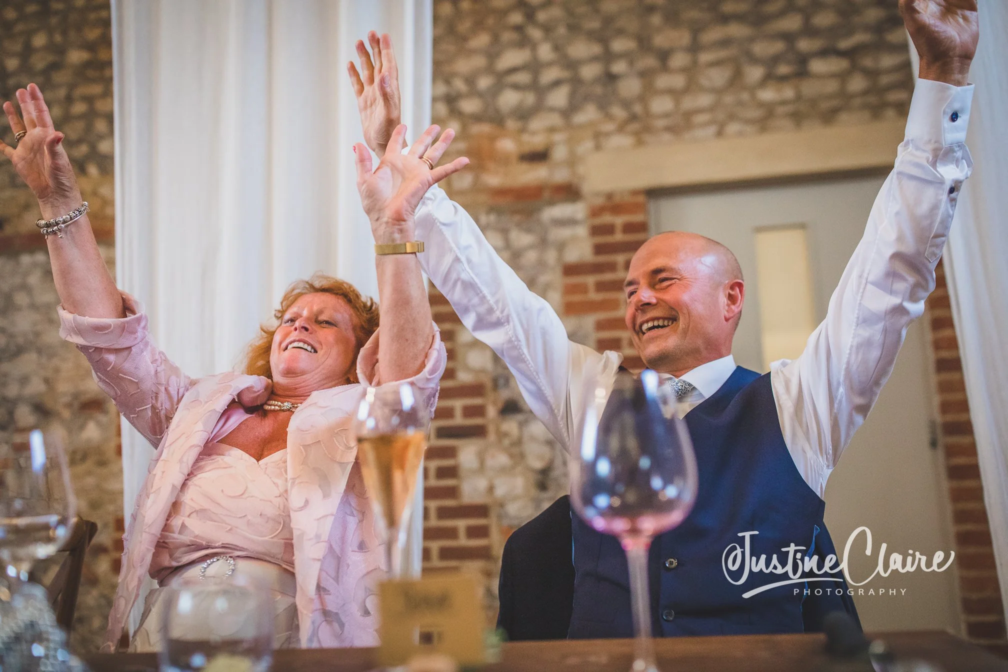 Two people, a woman and a man, sitting at a table with drinks, raising their hands and smiling joyfully during a celebration or toast, with a rustic brick wall in the background.