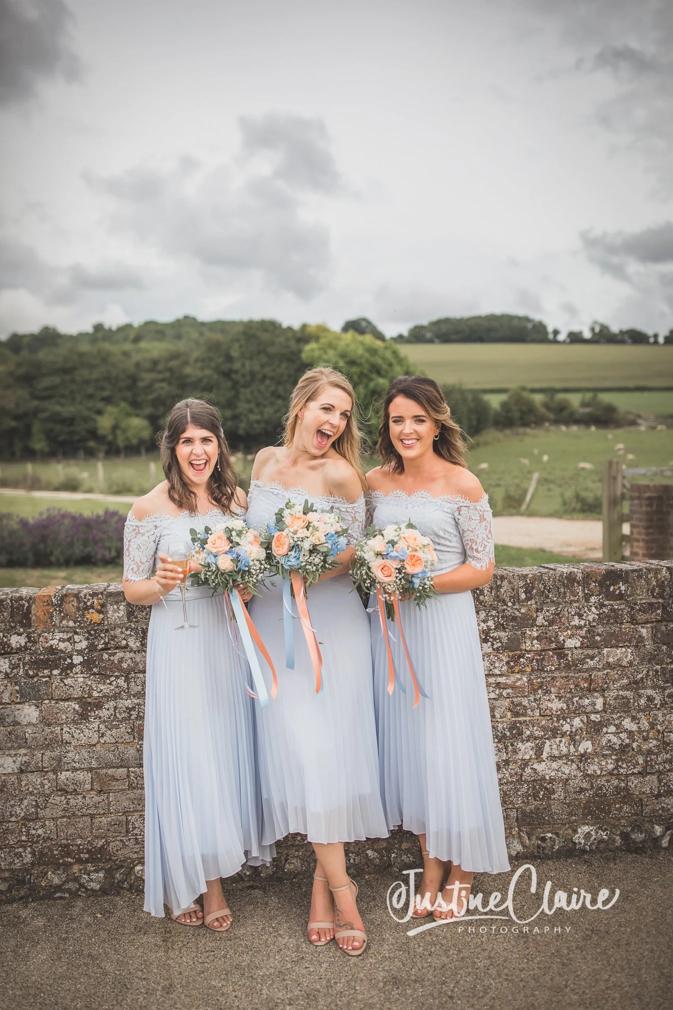 Three women in light blue bridesmaid dresses standing outdoors, holding bouquets, smiling and laughing.