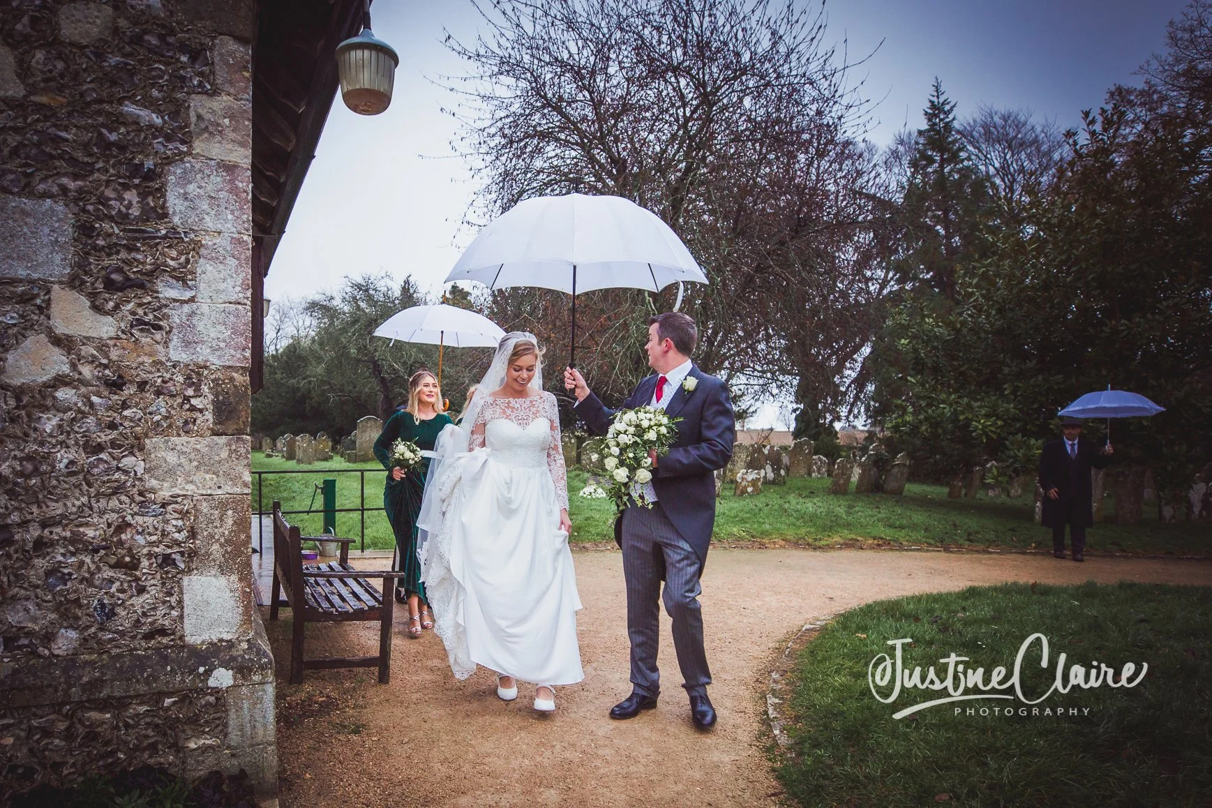 Wedding couple walking outdoors in rain, with bridesmaid holding bouquet behind them, bride in white dress and veil, groom in suit with boutonniere, carrying bouquet, holding white umbrellas, stone building wall on left, trees and grass in background