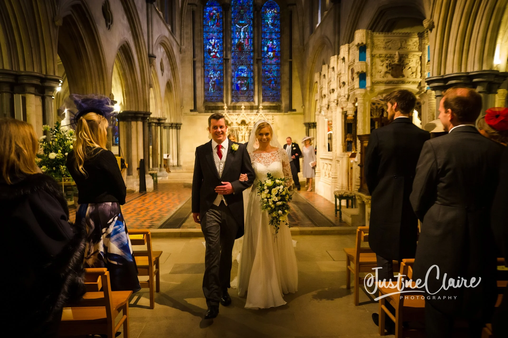 Bridal couple walking down the aisle in a church after their wedding ceremony, surrounded by guests seated on chairs, with stained glass windows in the background.