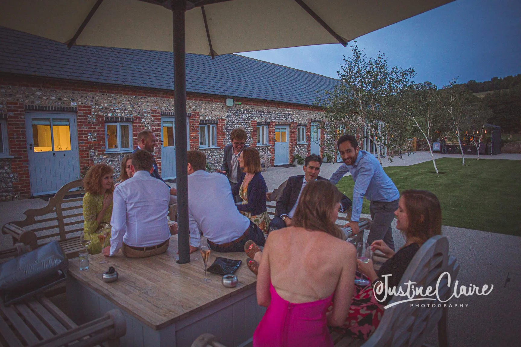 Group of people enjoying an outdoor dinner gathering at sunset, seated around a wooden table under a large umbrella, with a brick building and landscaped yard in the background.