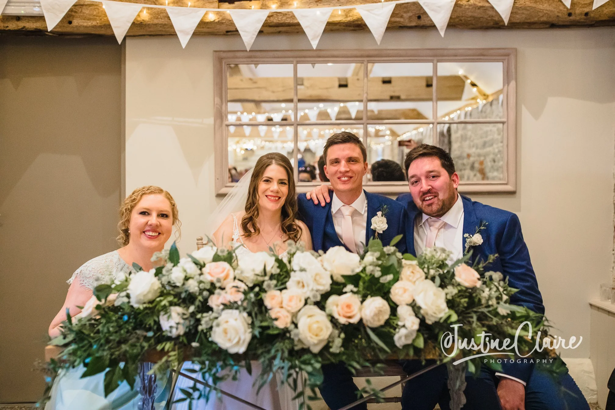 A group of four people, two women and two men, smiling and sitting behind a large floral arrangement at a wedding reception.