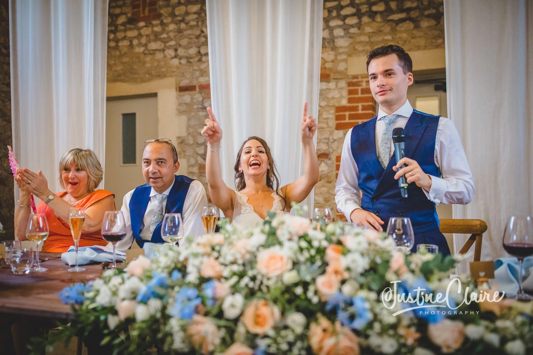 A wedding reception with four people seated at a table decorated with a floral centerpiece, with a woman in the middle raising her hands and smiling, a young man on the right holding a microphone, an older woman on the far left clapping, and a man ne