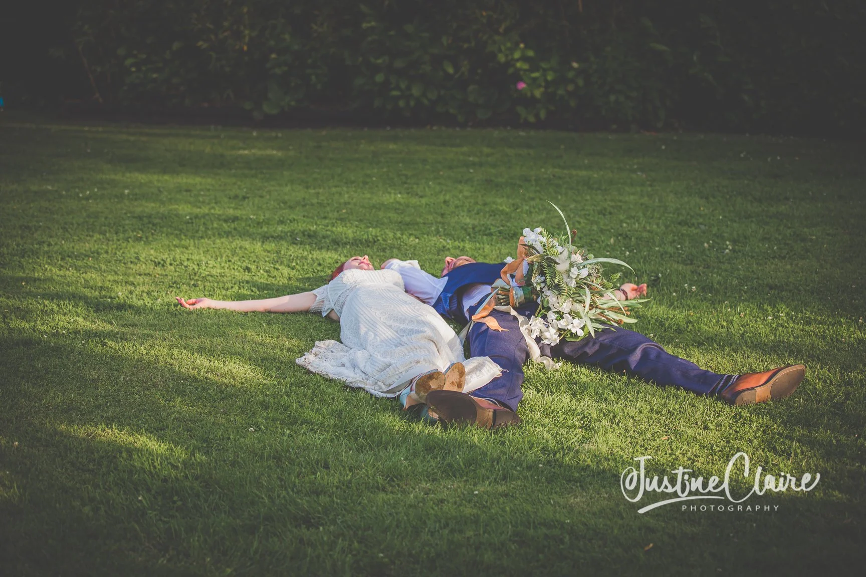 A couple lying on the grass after their wedding, with the bride's head resting on the groom's shoulder, surrounded by flowers.