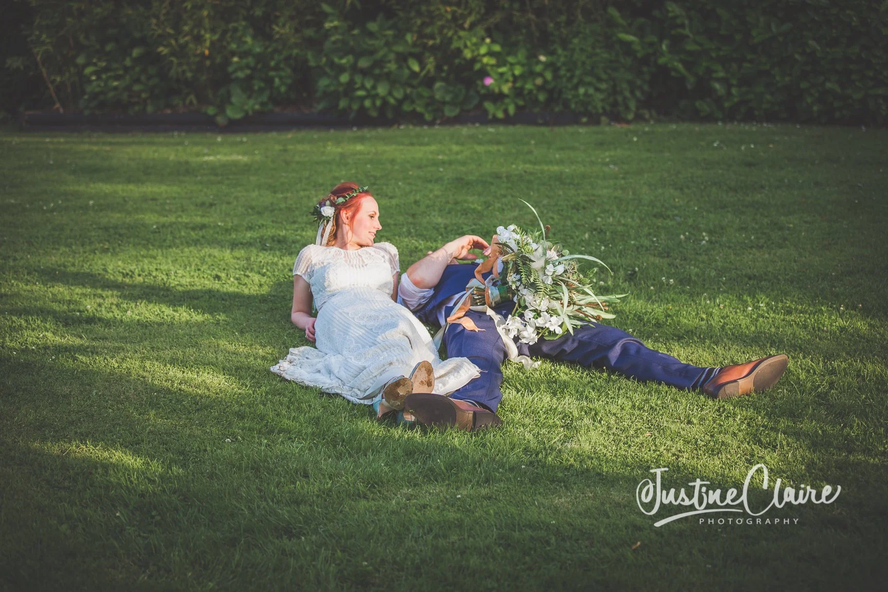 A bride and groom lying on the grass during their wedding, with the bride in a white dress and floral crown, and the groom holding a large bouquet of flowers.
