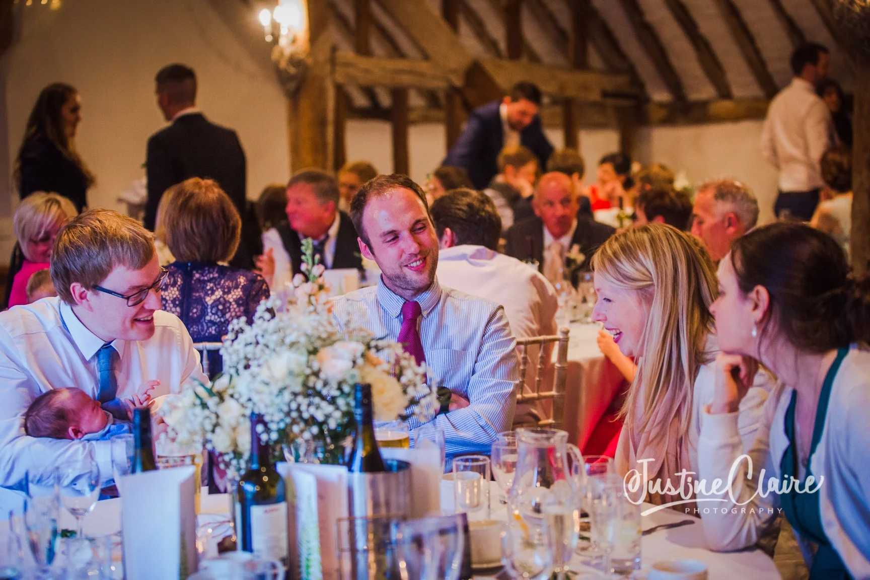 Guests seated at a wedding reception table, smiling and talking, with a centerpiece of white flowers and bottles of beverages, in a rustic venue with wooden beams.