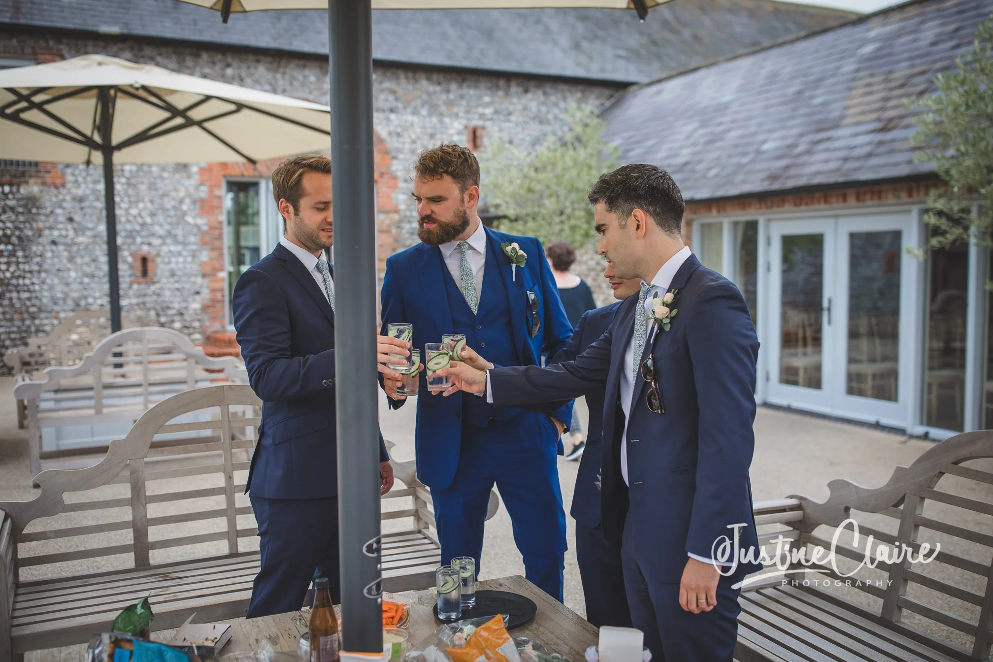 Four men in suits toasting drinks at an outdoor gathering, with a table of snacks and glasses, in a backyard setting with a brick and stone house.