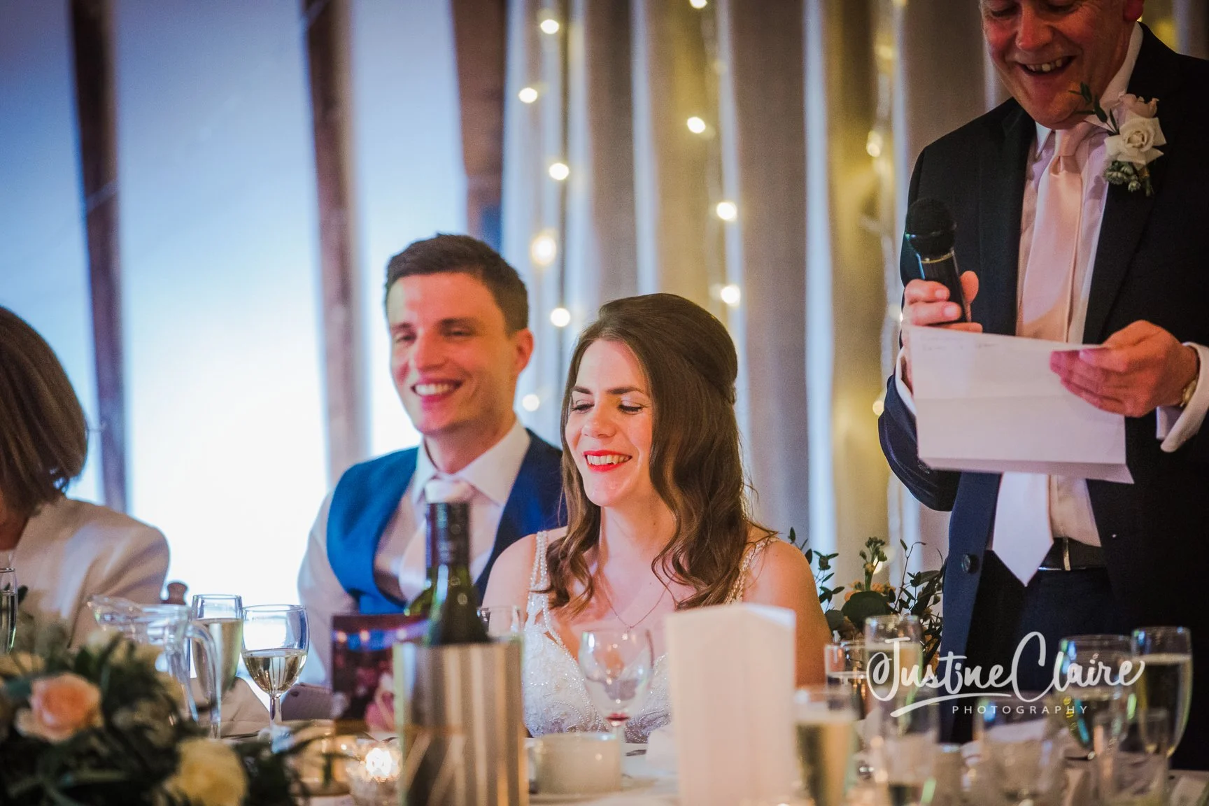 A happy bride smiling during her wedding reception seated at a decorated table with drinks, with a man in a blue suit smiling beside her, and a man in a black suit giving a speech while holding a microphone and notes, under warm string lights.