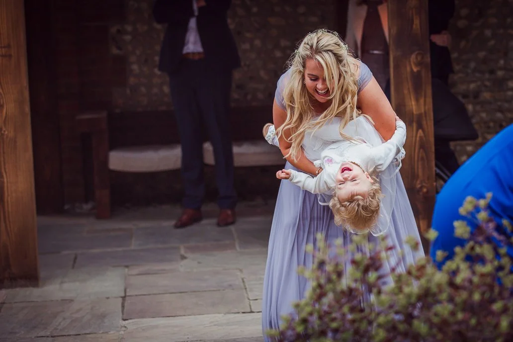 A woman in a light blue dress is happily swinging a laughing young girl by her arms at a celebration or wedding reception, with a man in the background and blurred flowers in the foreground.
