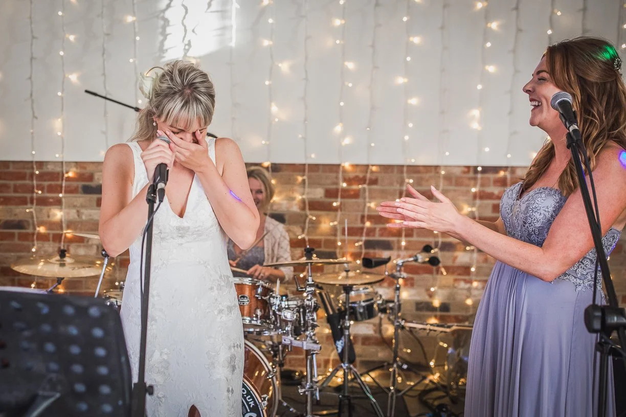 A woman in a white dress is crying and covering her face with her hands while holding a microphone. Another woman in a lavender dress is clapping and smiling. A drummer is playing in the background, with fairy lights and a brick wall behind them.