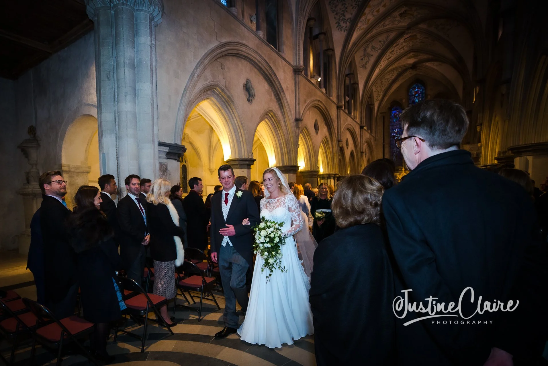 A newlywed bride and groom walking arm in arm down the aisle in a church, surrounded by guests. The bride is holding a large bouquet of white flowers and wearing a white lace dress and veil. The groom is dressed in a dark suit with a red tie. The chu
