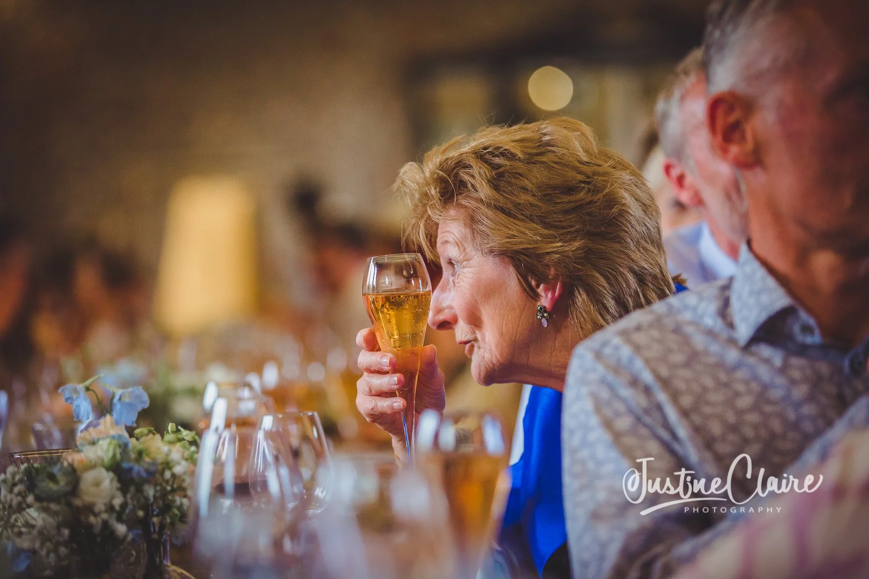 An older woman with short, curly hair holds a glass of champagne close to her face at a formal dinner, sitting beside a man with gray hair. The table is decorated with flowers and glassware, and other guests are visible in the blurred background.