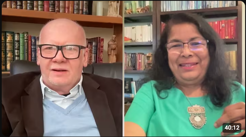 Two people engaging in a video call, sitting in rooms with bookshelves in the background. One person is wearing glasses and a suit, the other is wearing a green top with a large pendant.