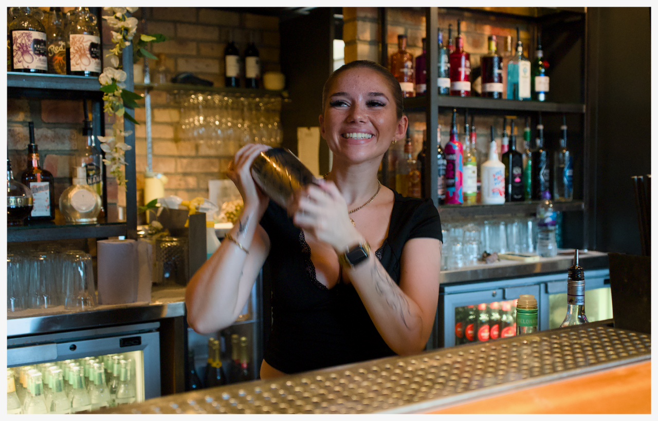 A woman with long blonde hair wearing earrings and a white spaghetti strap top, holding a cocktail shaker, behind a bar with liquor bottles.
