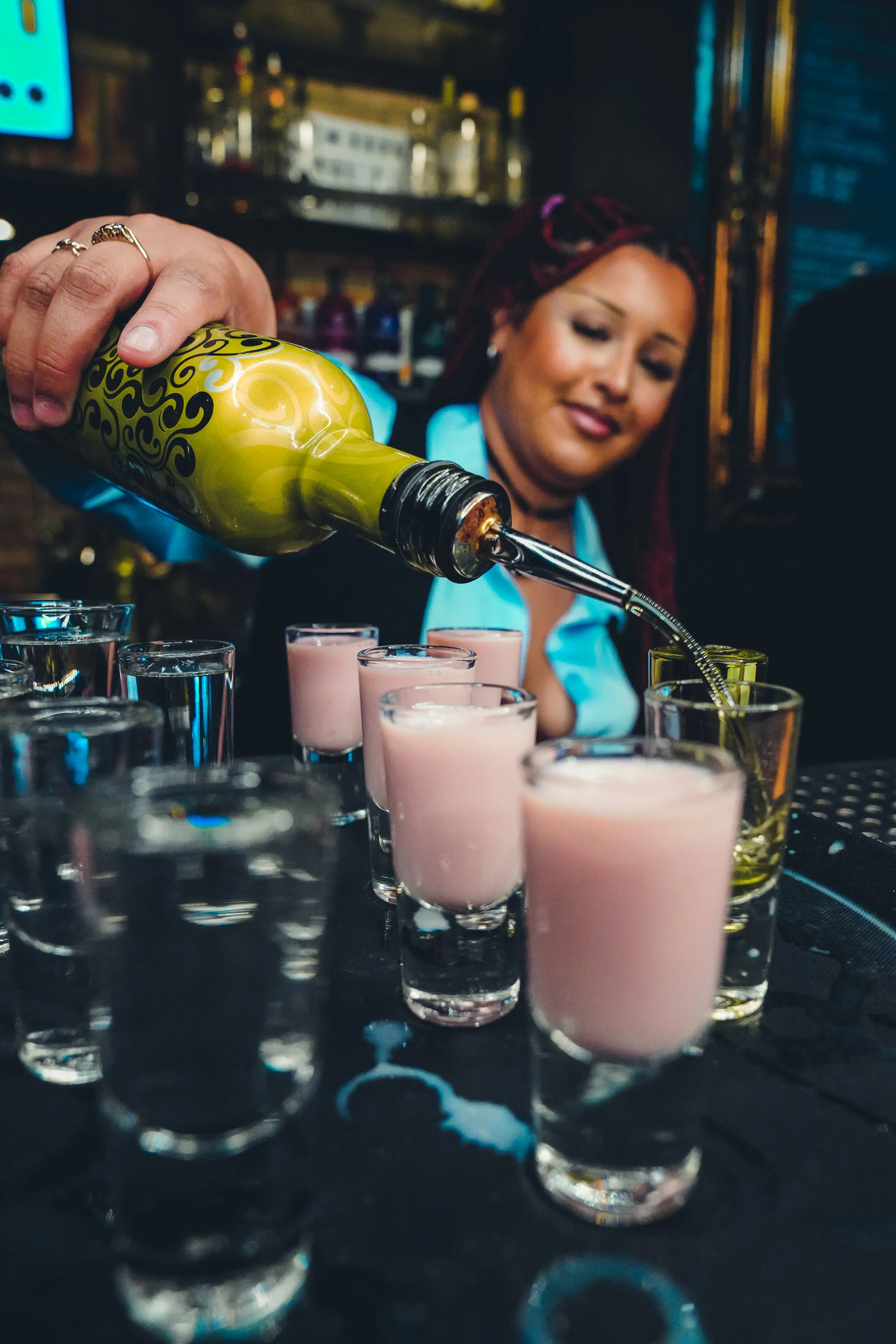 Group of ten women in a bar or club, some taking shots and others holding drinks, with pink and purple lighting.