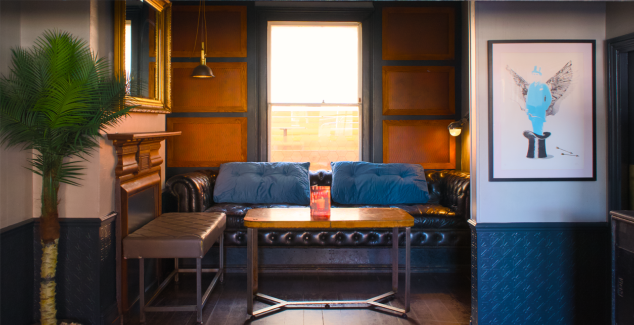 Cozy indoor setting with a black leather sofa, two blue pillows, a small brown table with a menu on it, a bench to the left, a large painting with a person with butterfly wings on the right, and a large leafed plant on the far left.