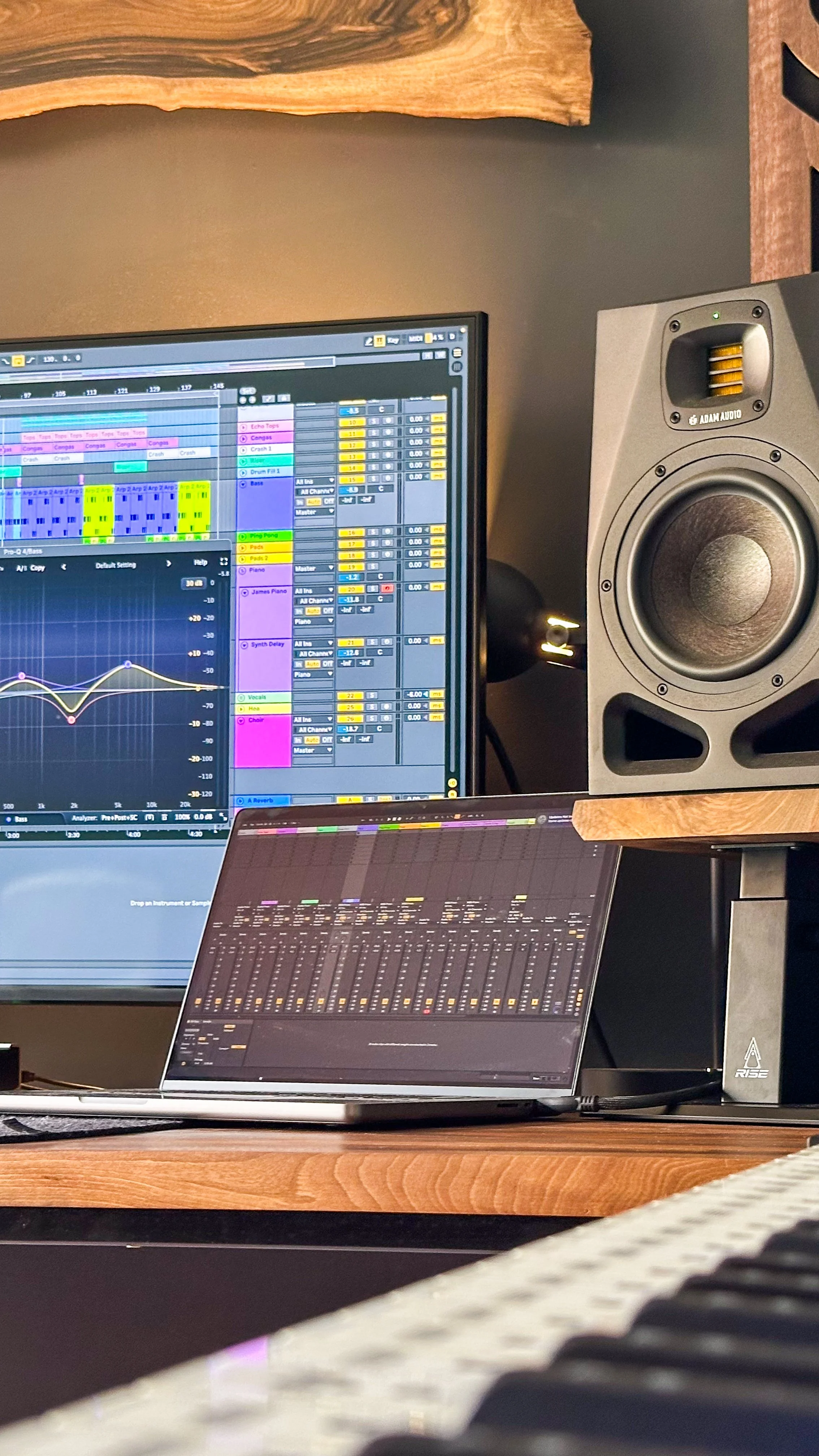 Studio speaker on a Rise Audio walnut monitor stand beside production screens and a laptop