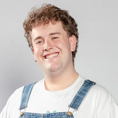 Een lachende jonge man met kort bruin haar voor een kleurrijke muurschildering op een gele muur.