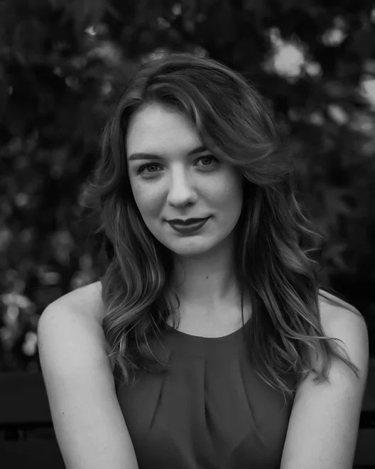 Black and white portrait of a young woman with wavy hair, wearing a sleeveless top, outdoors with trees in the background, smiling slightly.