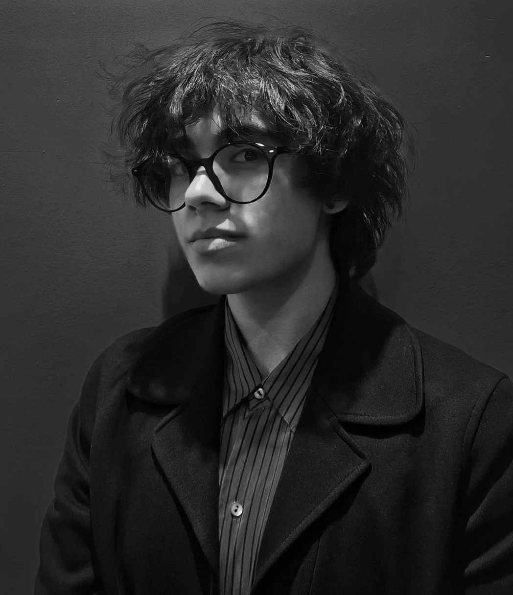 Black-and-white photo of a young person with curly hair and glasses, wearing a blazer and a striped shirt, standing against a dark wall.