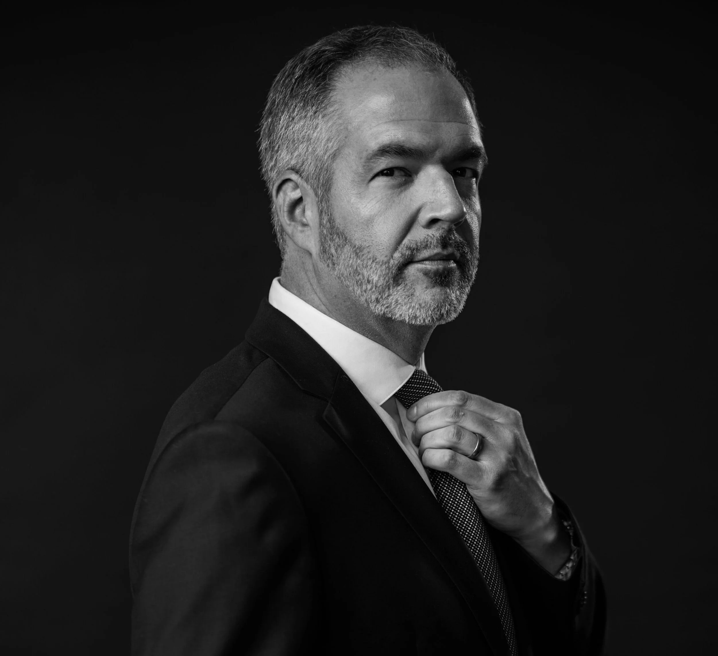 Black and white portrait of a middle-aged man with styled hair and a beard, wearing a suit and tie, adjusting his collar against a dark background.