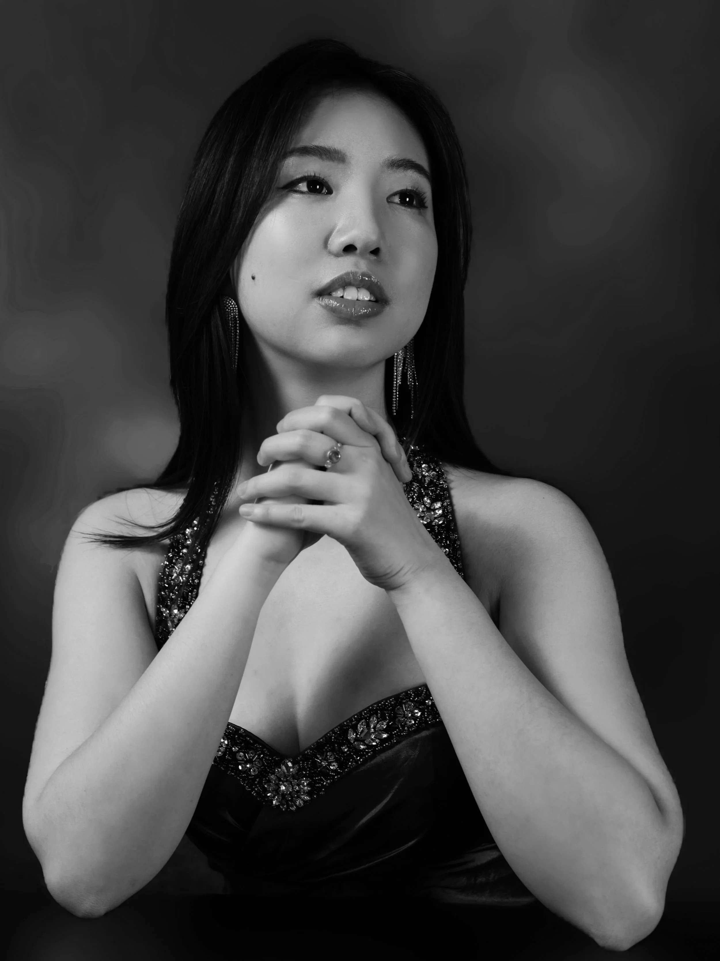 Black and white portrait of a woman with long dark hair, wearing a beaded dress and jewelry, with hands clasped near her chin, looking slightly to the side.