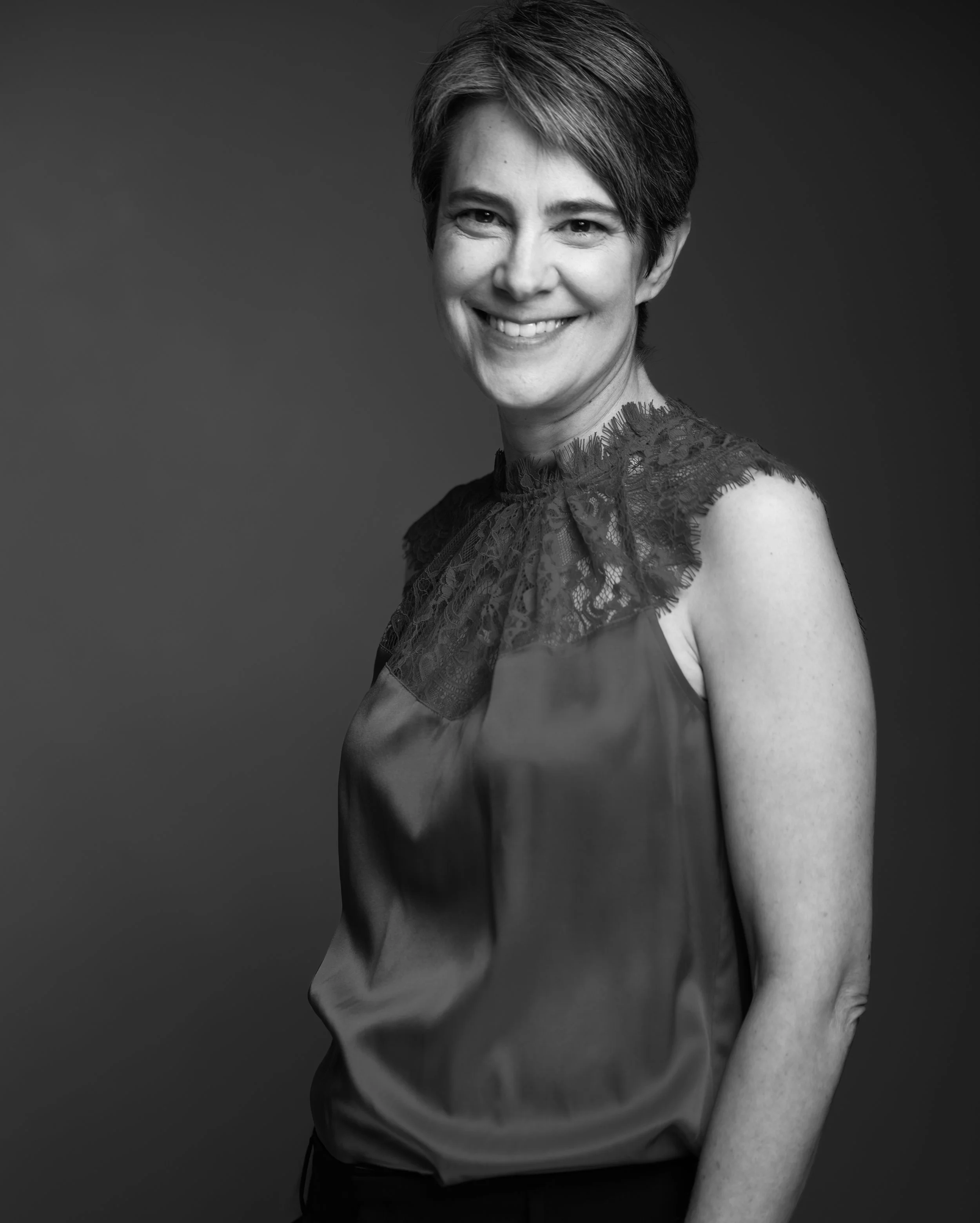 Black and white portrait of a smiling woman with short hair, wearing a satin blouse with lace details on the shoulders.
