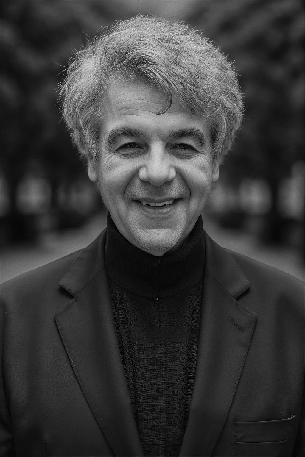 Black and white portrait of a smiling man with wavy gray hair, wearing a dark blazer and turtleneck sweater, outdoors with blurred trees in the background.