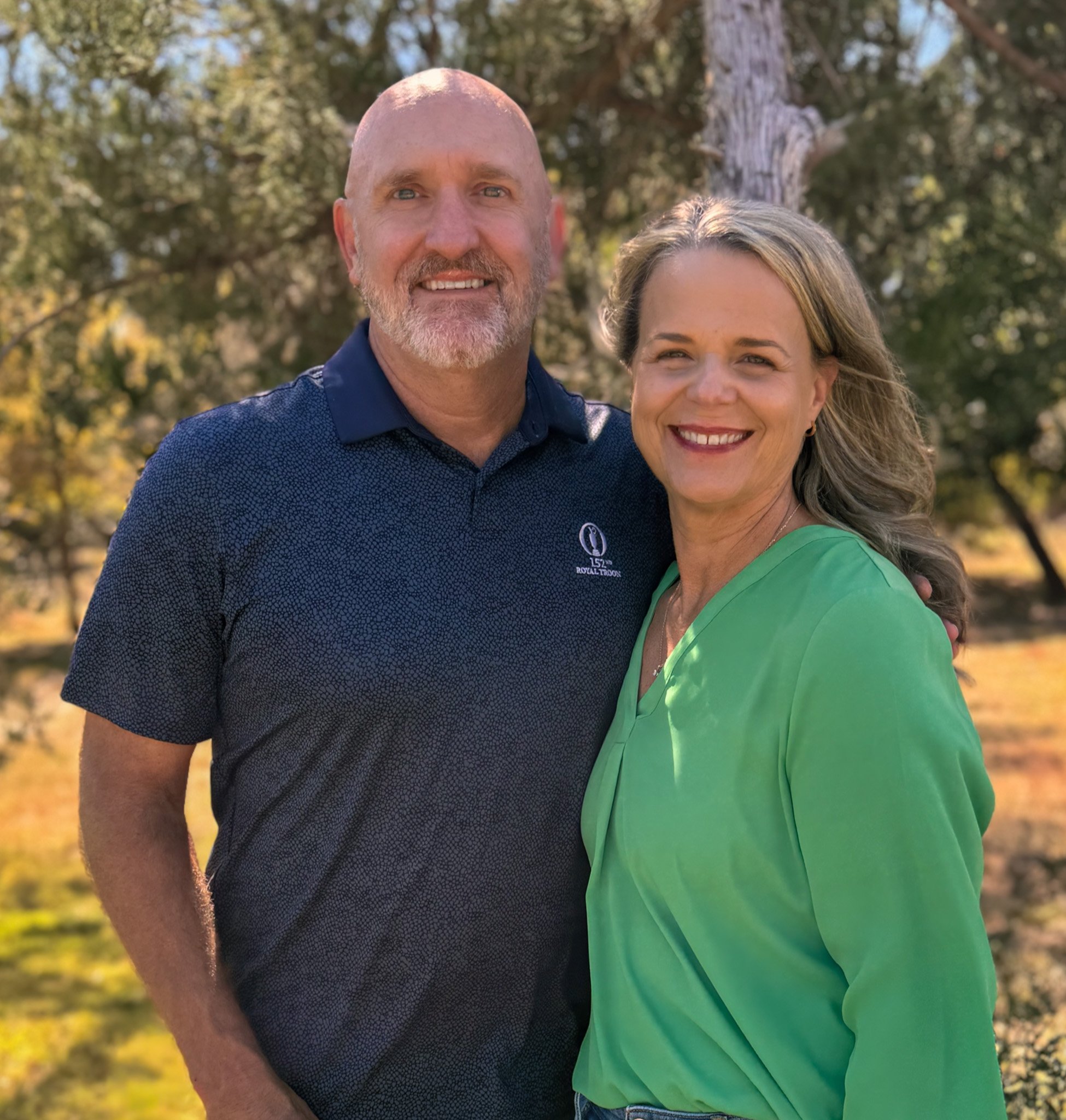 A smiling man and woman outdoors with trees in the background, the man wearing a blue shirt and the woman wearing a green top.