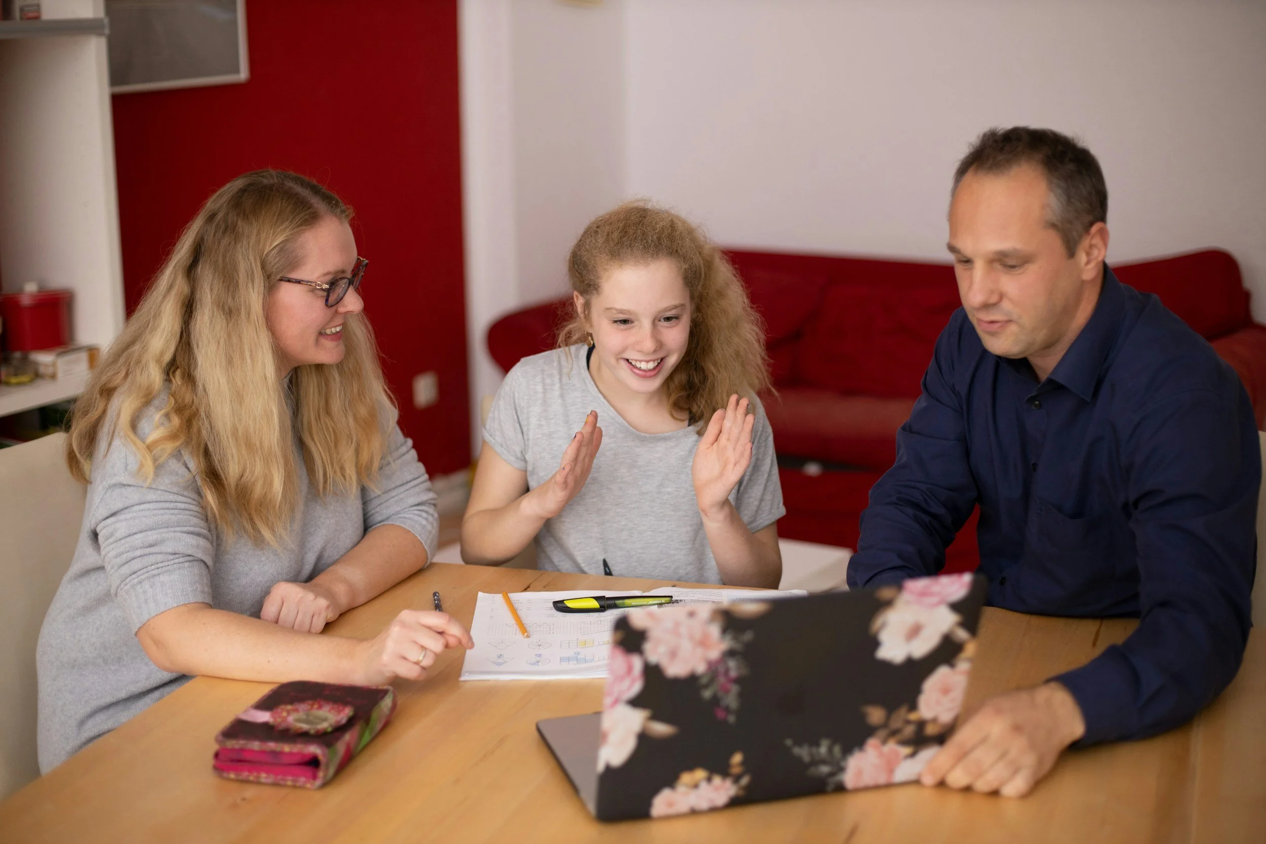 A family sitting at a table, smiling and engaging with each other, with notebooks, pens, and a floral-patterned laptop cover visible.