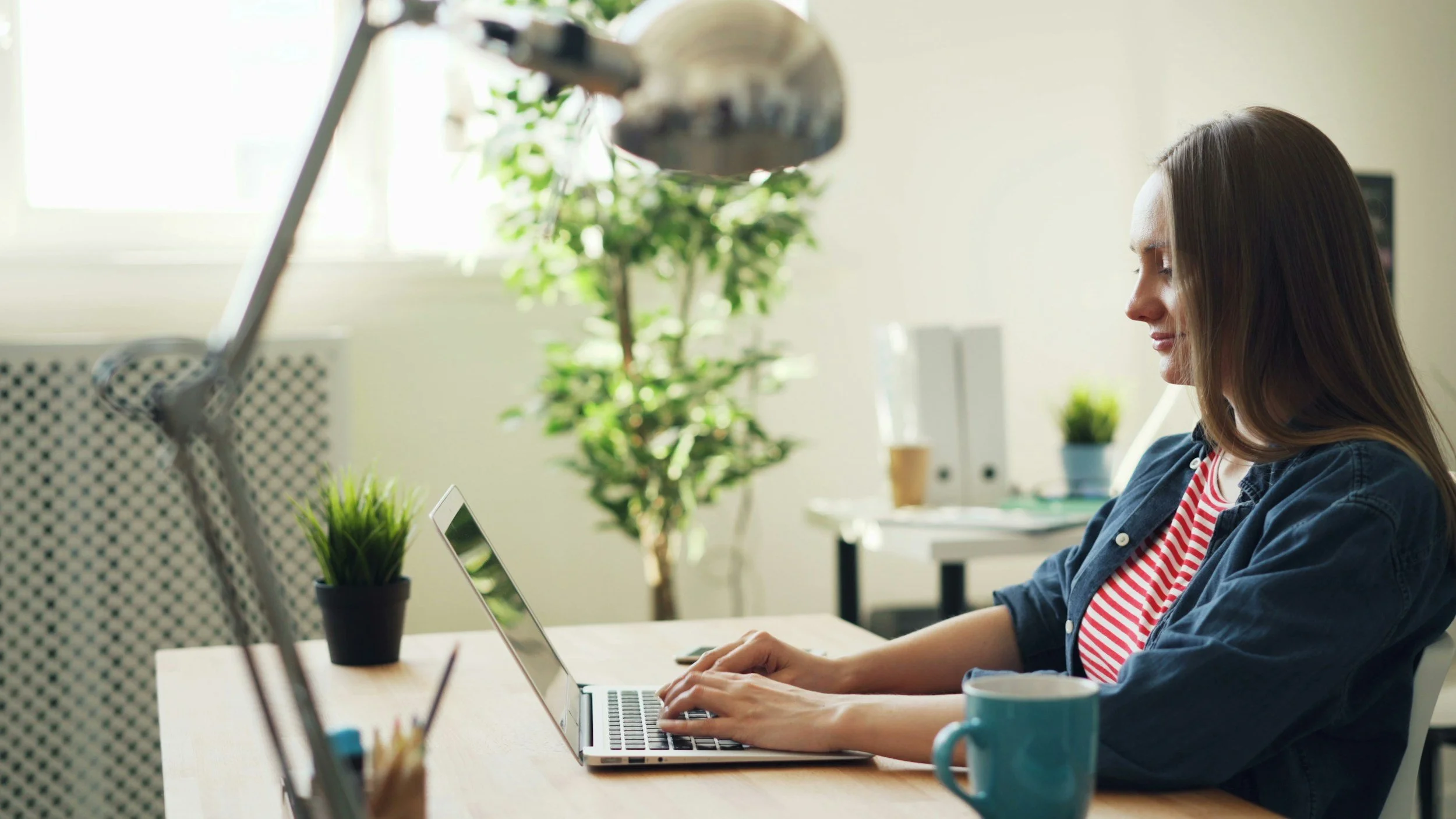 A woman with long brown hair, wearing a denim jacket over a red and white striped shirt, sitting at a desk and working on a laptop in a bright, plant-filled room.