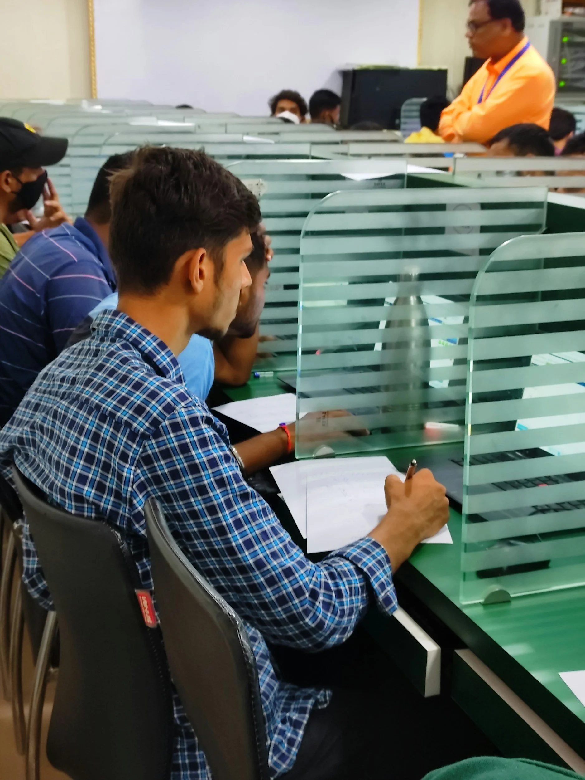 People sitting at desks divided by glass partitions in a classroom or training room, with an instructor standing at the front.