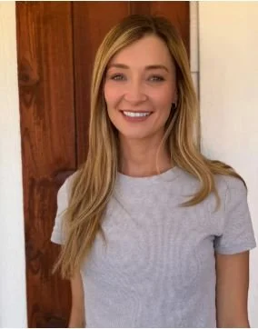 A young woman with long blonde hair smiling, wearing a light gray t-shirt, standing indoors with a wooden door in the background.